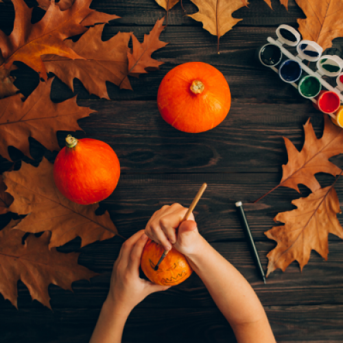 A table with pumpkins, autumn leaves, and paints; a person is painting a pumpkin with a brush, evoking a fall crafting vibe.