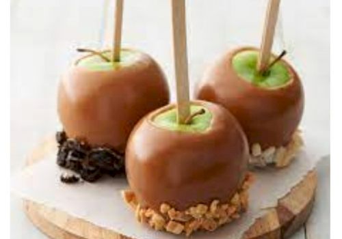 The image shows caramel apples on sticks, with toppings like nuts and cookie crumbs, placed on a wooden board.