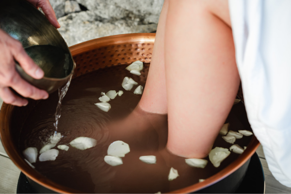 Feet soaking in a copper basin with water and rose petals, while additional water is being poured in.