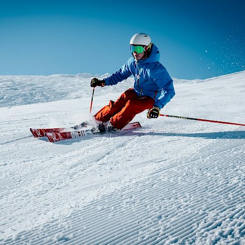 A skier in colorful gear is making a sharp turn on a sunny, snowy slope under a clear blue sky.