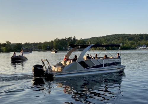 A pontoon boat with several people is on a calm body of water, with trees and another boat in the background, under a clear sky.