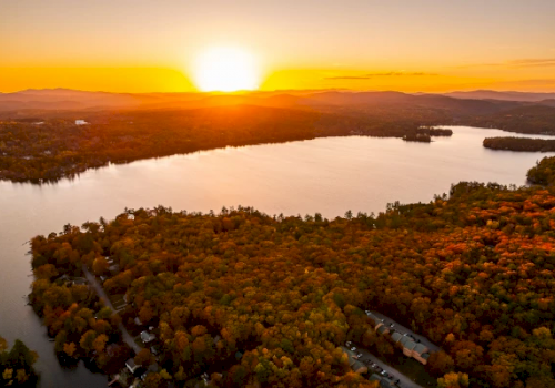 A scenic sunset over a lake surrounded by a forest with vibrant autumn foliage, creating a picturesque and serene landscape.