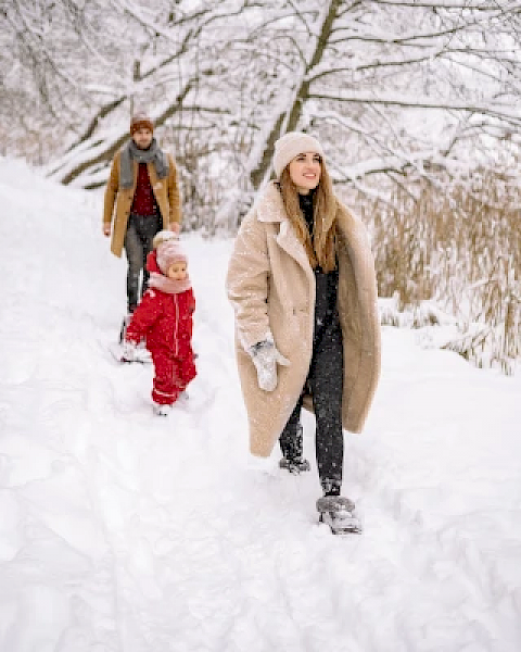 A family walking through a snowy path, with trees lining the background and bundled in winter clothing, enjoying a winter day outdoors.