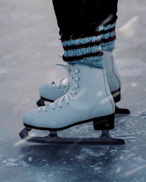 The image shows a close-up of someone's feet wearing white ice skates on an ice rink, with snow falling lightly around them.