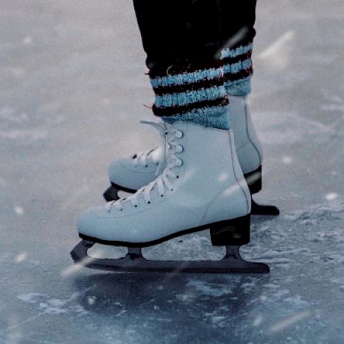 The image shows a close-up of someone's feet wearing white ice skates on an ice rink, with snow falling lightly around them.