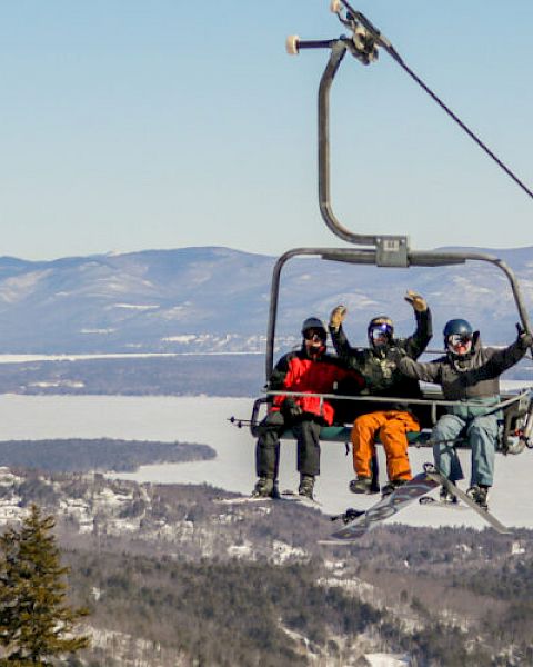 People are on a ski lift, enjoying scenic mountain views with snowy landscapes and evergreen trees in the background.