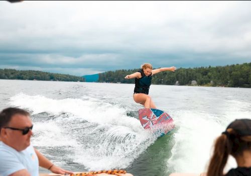 A person is wakeboarding on a lake, performing a trick on a wave behind a boat, with two people watching in the foreground.