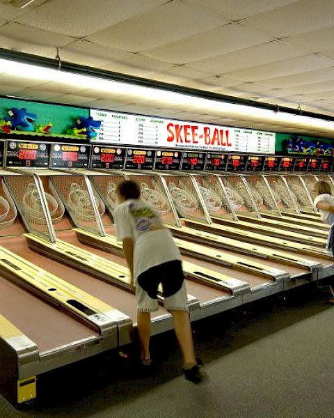 People playing skee-ball in an arcade lined with multiple alleys and a colorful sign above the lanes.