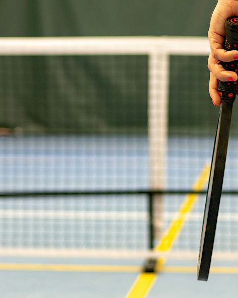 A hand holding a pickleball paddle and a yellow pickleball on an indoor court with a net in the background.