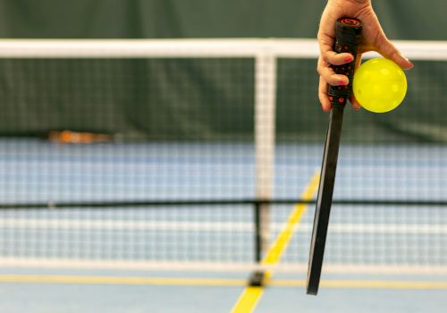 A hand holding a pickleball paddle and a yellow pickleball on an indoor court with a net in the background.