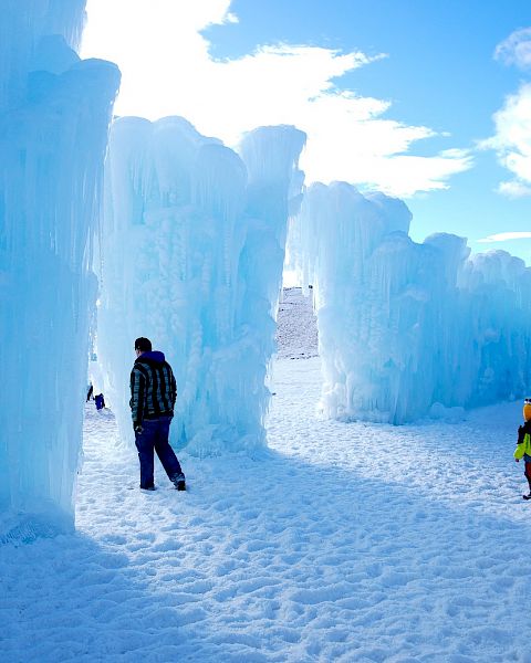 The image shows people walking through large ice structures under a bright blue sky, with snow covering the ground.