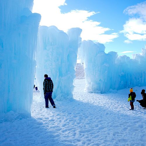 The image shows people walking through large ice structures under a bright blue sky, with snow covering the ground.