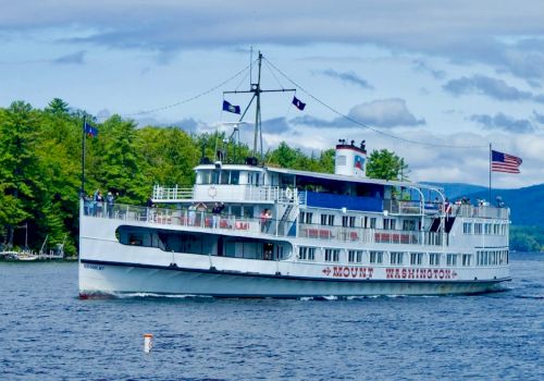 A white boat named Mount Washington cruises on a lake, with a U.S. flag atop. Tree-lined shore and hills are visible in the background.