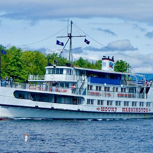 A white boat named Mount Washington cruises on a lake, with a U.S. flag atop. Tree-lined shore and hills are visible in the background.