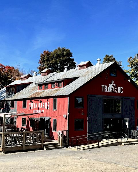 A red barn-like brewery building surrounded by trees, with outdoor seating and clear blue skies above.