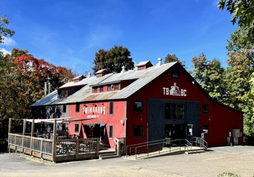 A red barn-like brewery building surrounded by trees, with outdoor seating and clear blue skies above.