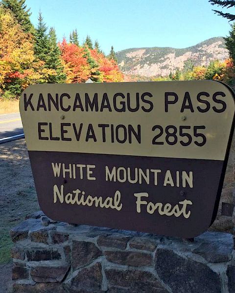 A sign for Kancamagus Pass, elevation 2855, in White Mountain National Forest with a road and colorful autumn trees in the background.