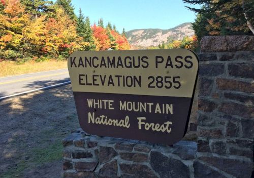 A sign for Kancamagus Pass, elevation 2855, in White Mountain National Forest with a road and colorful autumn trees in the background.