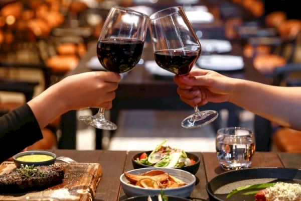 Two people clink wine glasses over a table with dishes, including a steak and salads, in a cozy restaurant setting.