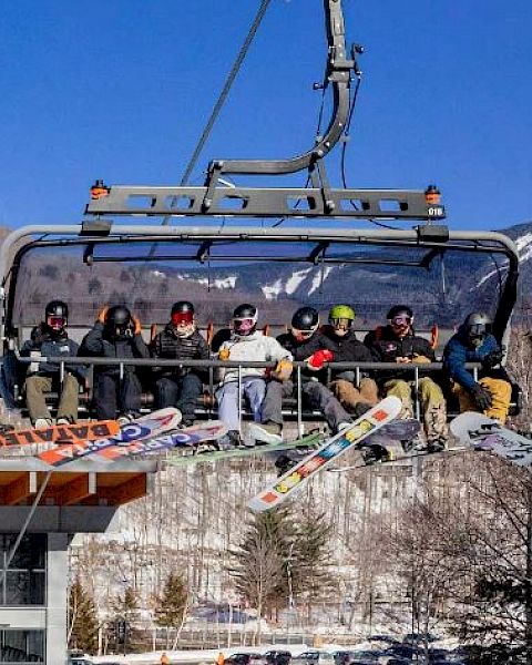 A group of people on a chairlift with snowboards in a mountainous, snowy area, enjoying a sunny day outdoors.