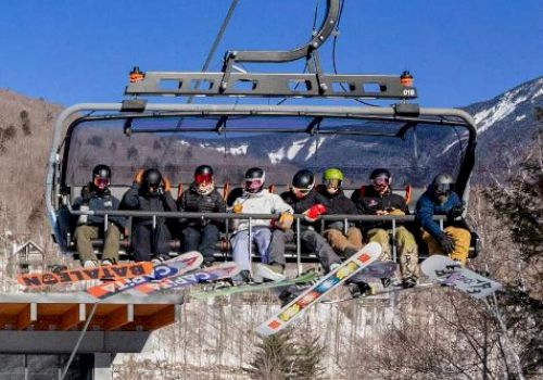 A group of people on a chairlift with snowboards in a mountainous, snowy area, enjoying a sunny day outdoors.