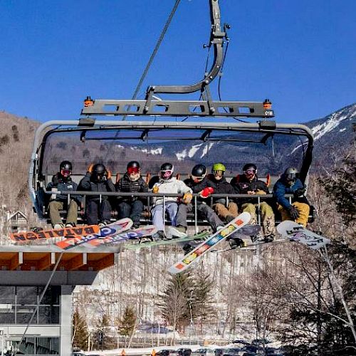 A group of people on a chairlift with snowboards in a mountainous, snowy area, enjoying a sunny day outdoors.