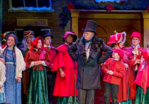 A group of people in period costumes on stage, possibly from a holiday-themed play, with festive decorations in the background.