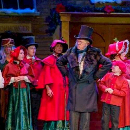 A group of people in period costumes on stage, possibly from a holiday-themed play, with festive decorations in the background.