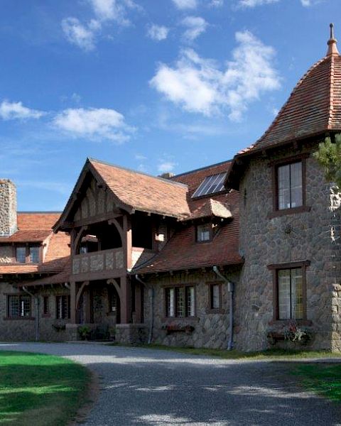 A large stone house with a rustic design, featuring steep roofs and a tower, surrounded by greenery and a clear blue sky.