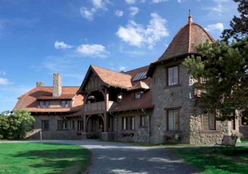 A large stone house with a rustic design, featuring steep roofs and a tower, surrounded by greenery and a clear blue sky.