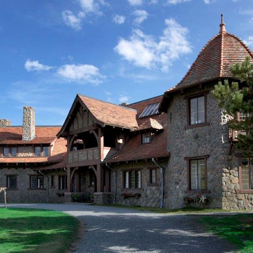 A large stone house with a rustic design, featuring steep roofs and a tower, surrounded by greenery and a clear blue sky.