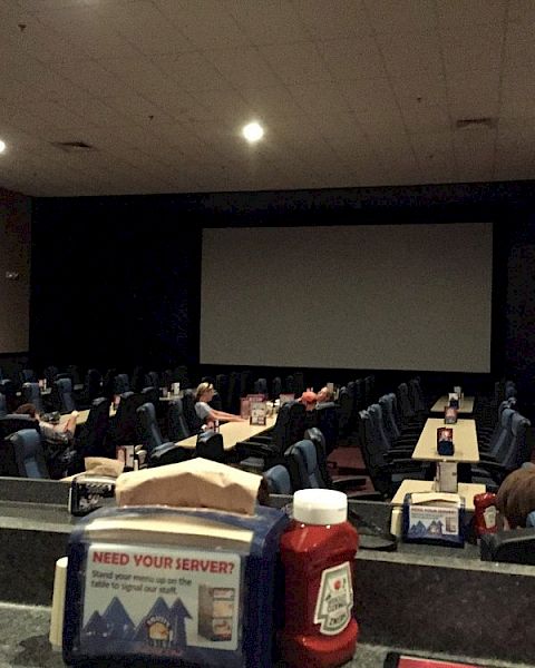 A dimly lit movie theater with tables and ketchup bottles, featuring a large screen and few people seated.