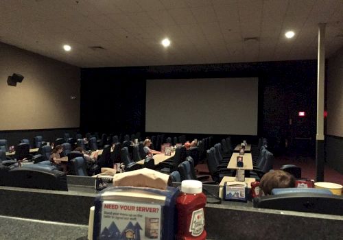 A dimly lit movie theater with tables and ketchup bottles, featuring a large screen and few people seated.