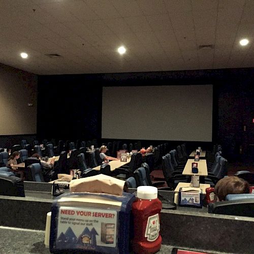 A dimly lit movie theater with tables and ketchup bottles, featuring a large screen and few people seated.