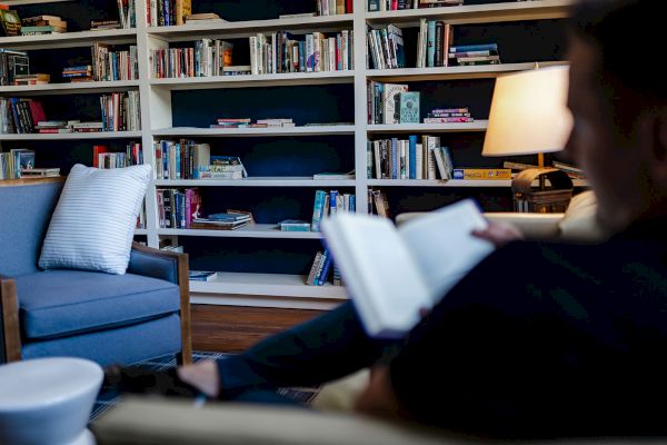 A person is reading a book in a cozy room with bookshelves, a lamp, and a chair with a pillow, creating a relaxing atmosphere.