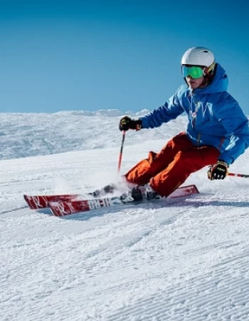 A skier in a blue jacket and red pants is carving down a snowy slope under a clear blue sky, wearing a helmet and goggles.