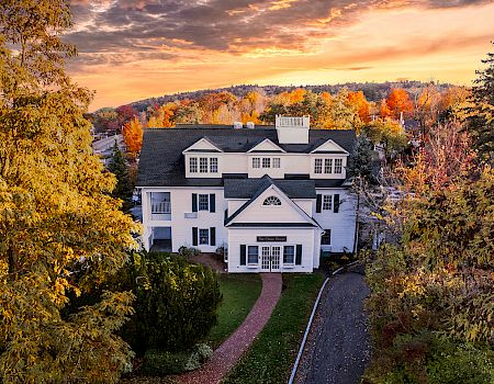 A large white house surrounded by colorful autumn trees, set under a dramatic sunset sky, with a brick path leading up to the entrance.