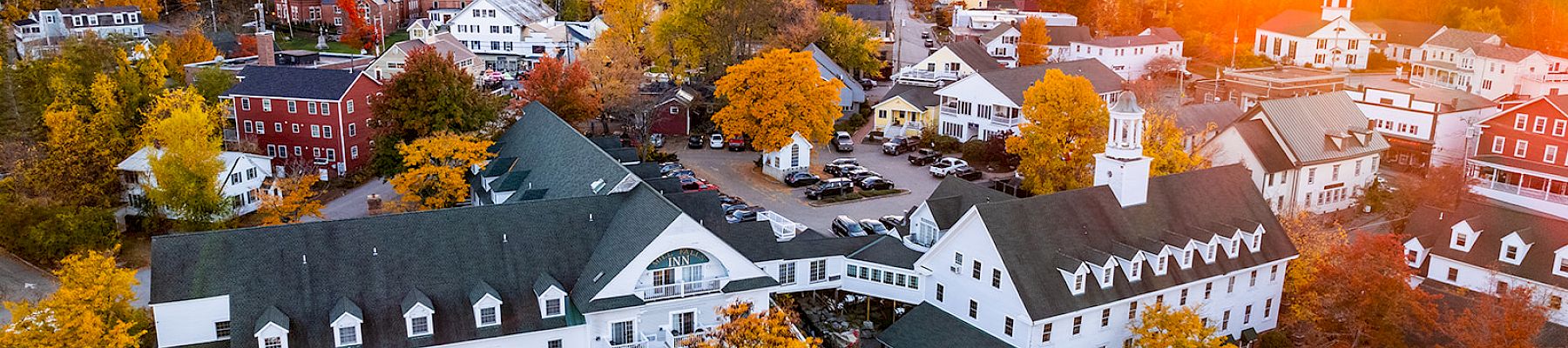 Aerial view of a quaint town at sunset, featuring colorful autumn trees, white buildings, and a church with a spire in the distance.