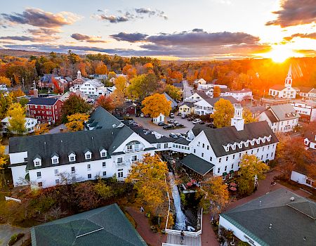 Aerial view of a quaint town at sunset, featuring colorful autumn trees, white buildings, and a church with a spire in the distance.